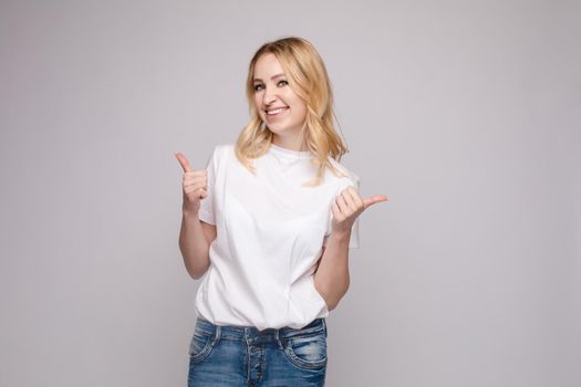 Front view of positive blonde wearing white shirt and jeans looking at camera and showing thumbs up. Young woman posing and laughing on grey isolated background. Concept of happiness.