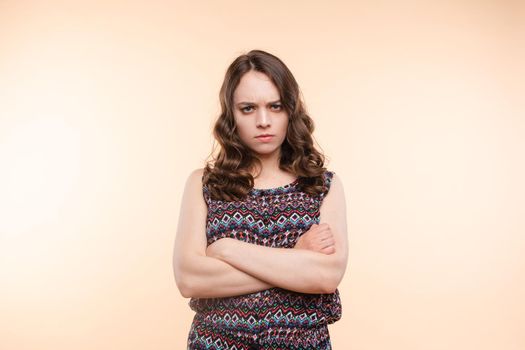 Studio portrait of unsatisfied young brunette caucasian woman with wavy hair in overall with colorful pattern holding arms folded and looking at camera with grief, dissatisfaction, anger and disbelief. Isolate.