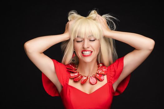 Fashion stressed blonde woman in red dress shout with open mouth holding head by hand medium closeup. Attractive worried female model posing having negative emotion isolated at black studio background