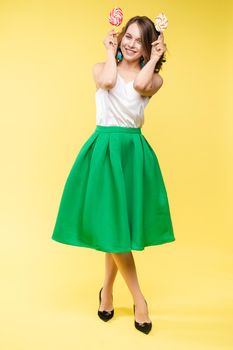 Full length studio portrait of laughing model in white top and green skirt and heels holding two sweet candies on her eyes like sunglasses with her mouth open. Isolate on yellow.