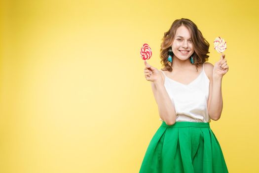 Full length studio portrait of laughing model in white top and green skirt and heels holding two sweet candies on her eyes like sunglasses with her mouth open. Isolate on yellow.