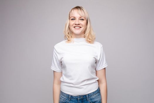 View from front of beautiful slim woman wearing white shirt and jeans standing steady on frey isolated background. Young blonde looking at camera, smiling and posing in studio.