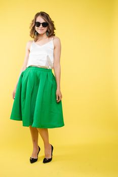 Summer outfit on orange background. Stylish and trendy beautiful young girl with wavy middle-length hair in white top and green bottom wearing trendy earrings and black sunglasses. She is smiling over orange background. Copyspace. Isolate.