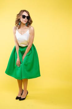 Summer outfit on orange background. Stylish and trendy beautiful young girl with wavy middle-length hair in white top and green bottom wearing trendy earrings and black sunglasses. She is smiling over orange background. Copyspace. Isolate.