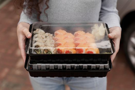 Close-up sushi in box healthy food delivery online service. Girl holds 2 sushi sets in hands. Japanese cuisine: rolls, soy sauce, wasabi.