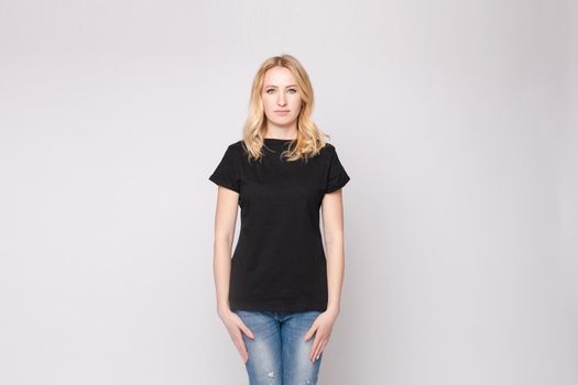 View from front of beautiful slim woman wearing black shirt and jeans standing steady on frey isolated background. Young blonde looking at camera, smiling and posing in studio.