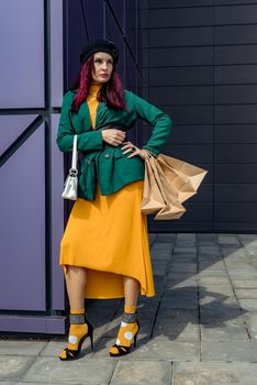 A happy shopaholic girl keeps her bags near the shopping center. A woman near the store is happy with her purchases, holding bags. She is wearing a yellow dress and a green jacket. Consumer concept