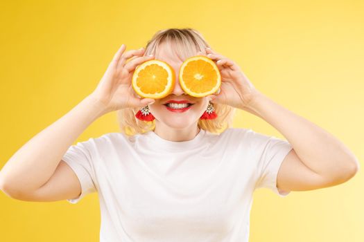 Front view of cheerful young blonde posing with fresh oranges on isolated background. Funny girl keeping fruit and closing eyes in studio. Concept of happiness and health.