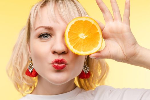 Studio headshot of young funny brunette with hairstyle and red lips holding halved oranges on eyes against bright yellow background.