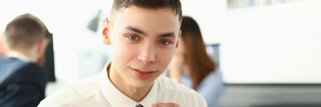 Portrait of unexperienced employee looking at camera with big joy. Man in classy tie sitting in big modern office with colleagues talking about important business project. Company meeting concept