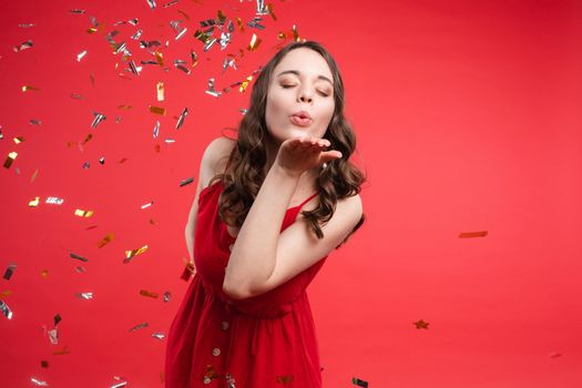 Front view of pretty brunette wearing red dress looking at camera while sparkles falling around in studio. Beautiful woman with hairstyle posing on isolated background. Concept of holiday.