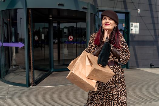 A happy shopaholic girl keeps her bags near the shopping center. A woman near the store is happy with her purchases, holding bags. Dressed in a leopard print dress. Consumer concept