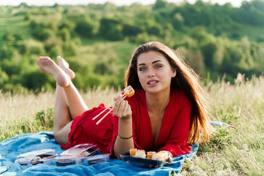 Attractive girl with 3 sushi sets lays on the blue cover on green hills background. Sexy girl in red dress gets sushi box from courier. Food delivery from restaurant