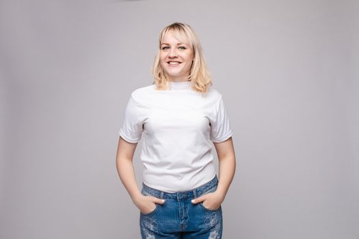 View from front of beautiful slim woman wearing white shirt and jeans standing steady on frey isolated background. Young blonde looking at camera, smiling and posing in studio.