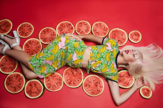 Sexy beautiful young woman lying on floor posing for photo shooting surrounded by watermelon full shot. Charming blonde female model in colorful clothing relaxing isolated at red studio background
