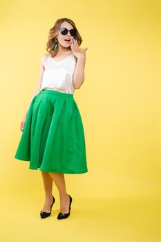 Summer outfit on orange background. Stylish and trendy beautiful young girl with wavy middle-length hair in white top and green bottom wearing trendy earrings and black sunglasses. She is smiling over orange background. Copyspace. Isolate.