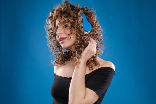 Front view of crazy curly woman with makeup wearing headphones and listening music on isolated background. Pretty girl dancing, singing and enjoying song in studio. Concept of disco and party.