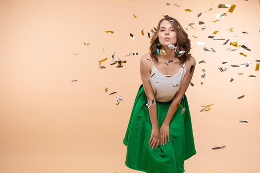 Happy funny woman rising up hand surrounded by falling shining multicolored confetti isolated at light studio background. Adorable smiling female enjoying fun having positive emotion medium long shot