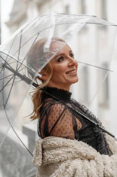 The blonde stands under a transparent umbrella during the rain. The fall season. Rear view. The woman is dressed in a black lace dress, her hair pulled back in a ponytail