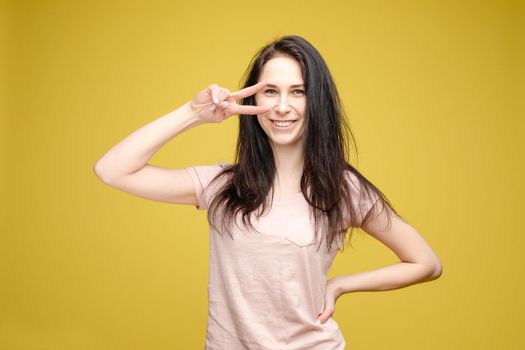 Studio portrait of charming brunette young woman in white top and green skirt holding peace sign near her eye. She is smiling at camera. Isolate on orange.