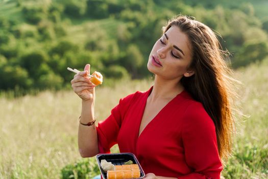 Sushi set and girl on green hills background. Food delivery service from japanese restaurant. Woman with blue eyes in red dress seats and eats sushi delivered by courier
