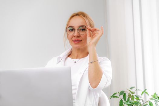 A blonde doctor in glasses or a cosmetologist in a white coat is sitting at a computer. She reads and types on the computer. Concept of medicine, beauty, cosmetologist, masseur