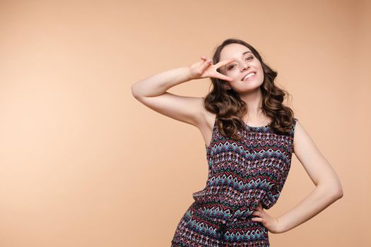 Studio portrait of charming brunette young woman in white top and green skirt holding peace sign near her eye. She is smiling at camera. Isolate on orange.