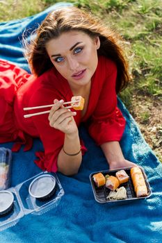 Attractive girl with 3 sushi sets lays on the blue cover on green hills background. Sexy girl in red dress gets sushi box from courier. Food delivery from restaurant