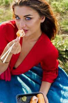 Sushi near lips of girl with blue eyes at picnic. Food delivery from japanese restaurant. Vertical photo advert for social networks. Pretty girl eats sushi set in the park.
