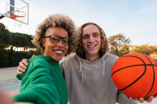 selfie of a latin woman and caucasian man smiling happy after playing in a city park basketball court, concept of friendship and urban sport in the street