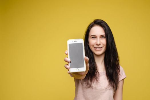 Studio portrait of amazed young caucasian woman with wavy hair wearing white top and green bottom holding mobile phone screen to camera. She is shocked or surprised with the news or info on the screen. Copyspace. Isolate on yellow.