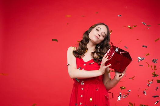 Portrait of adorable smiling young woman long curly hair posing isolated at red studio background. Beautiful happy female enjoying relaxing surrounded by shining colorful confetti looking at camera