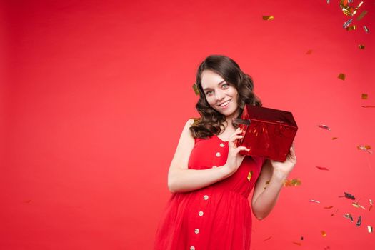 Front view of happy young girl wearing red dress looking at camera and twirling hair with hand in studio. Curly model posing and smiling on isolated background. Concept of emotions and celebration.