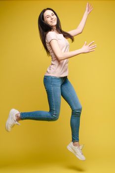 Full length isolate portrait of positive brunette young girl in jeans and t-shirt and sneakers jumping over yellow background. She is smiling at camera.