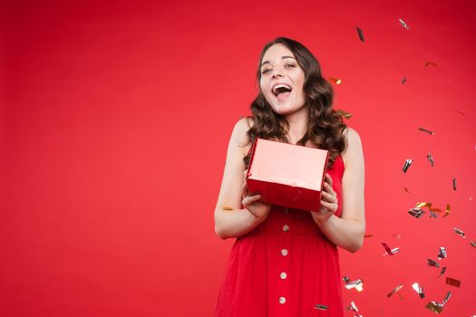 Front view of pretty brunette wearing red dress looking at camera while sparkles falling around in studio. Beautiful woman with hairstyle posing on isolated background. Concept of holiday.