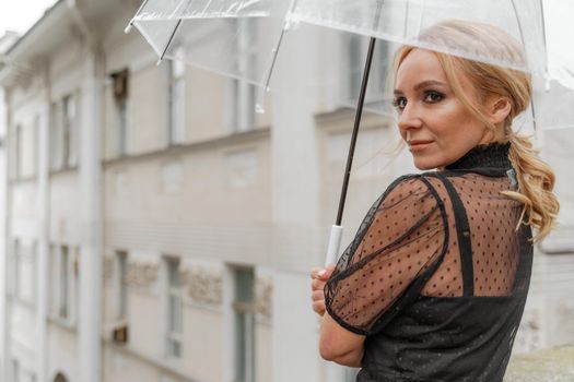 The blonde stands under a transparent umbrella during the rain. The fall season. Rear view. The woman is dressed in a black lace dress, her hair pulled back in a ponytail