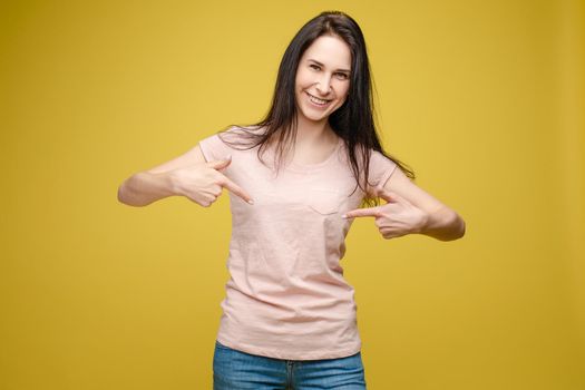 Cropped view from front of happy brunette standing in white shirt and jeans and pointing at outfit with fingers. Cheerful girl laughing and posing on grey isolated backround. Concept of casual style.