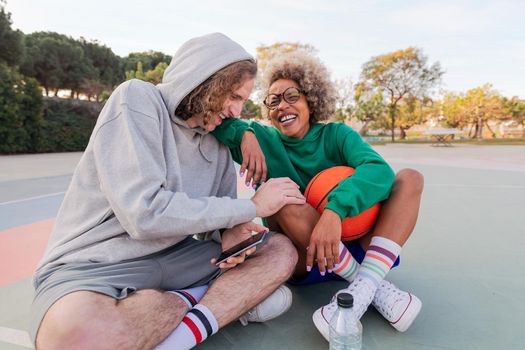man and woman laughing in amusement looking at things on their mobile phone sitting after playing basketball in a city park, concept of friendship and urban sport in the street