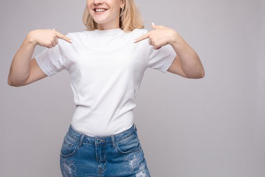 Cropped view from front of happy brunette standing in white shirt and jeans and pointing at outfit with fingers. Cheerful girl laughing and posing on grey isolated backround. Concept of casual style.