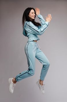 Happy brunette wearing blue tracksuit jumping and laughing on grey isolated background. Young cheerful woman with long hair having fun and posing in studio. Concept of happiness and motion.