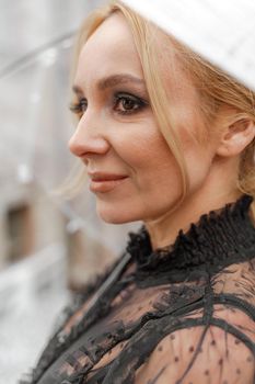 The blonde stands under a transparent umbrella during the rain. The fall season. Rear view. The woman is dressed in a black lace dress, her hair pulled back in a ponytail