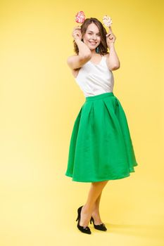 Full length studio portrait of laughing model in white top and green skirt and heels holding two sweet candies on her eyes like sunglasses with her mouth open. Isolate on yellow.