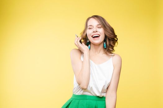 Studio portrait of amazed young caucasian woman with wavy hair wearing white top and green bottom holding mobile phone screen to camera. She is shocked or surprised with the news or info on the screen. Copyspace. Isolate on yellow.