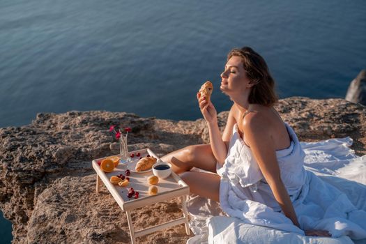 Woman covered with a blanket of bed relaxing and watching the seascape at sunrise. She holds a cup of coffee in her hand in front of her is a table with fruits and croissants. Wanderlust and freedom concept scene