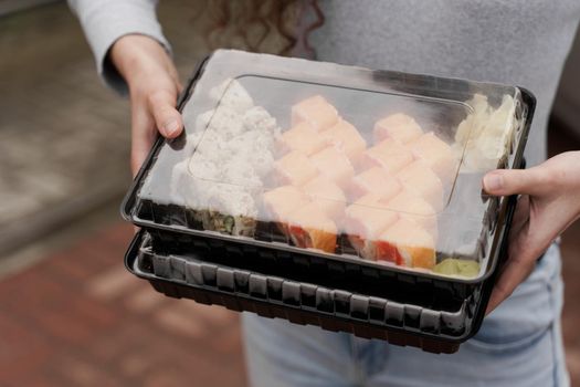 Close-up sushi in box healthy food delivery online service. Girl holds 2 sushi sets in hands. Japanese cuisine: rolls, soy sauce, wasabi.
