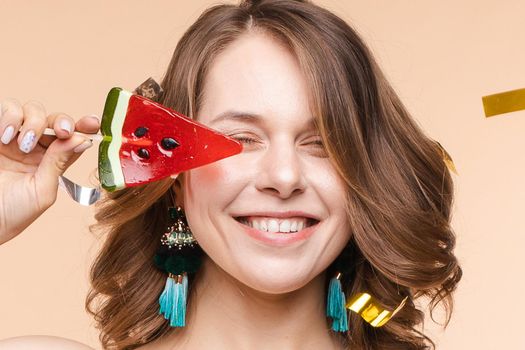 Playful elegant smiling woman posing with piece watermelon candy surrounded shining colorful confetti. Happy young female having positive emotion looking at camera isolated at light studio background