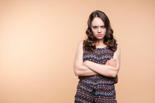 Studio portrait of unsatisfied young brunette caucasian woman with wavy hair in overall with colorful pattern holding arms folded and looking at camera with grief, dissatisfaction, anger and disbelief. Isolate.