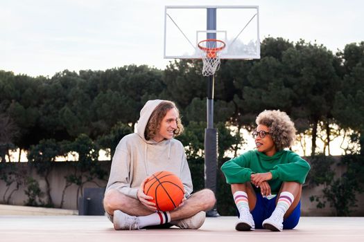 latin woman and caucasian man chatting sitting on the court after basketball practice, concept of friendship and urban sport in the street, copy space for text