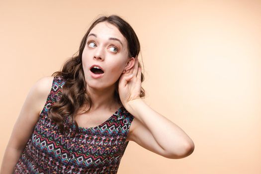 Studio portrait of curious brunette girl in multicolored top listening to the news or gossips with her ear.