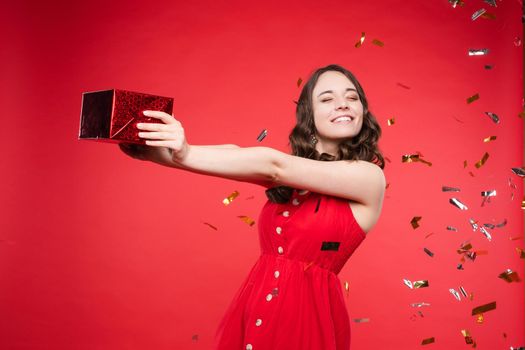 Studio portrait of beautiful young brunette lady with wavy hair in summer red dress with buttons smiling at camera. She is sprinkled or showered with sparkling confetti on red background. Isolate.